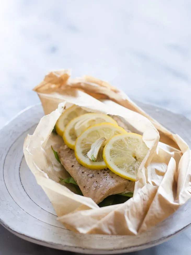 A salmon filet in a piece of parchment paper with snow peas underneath and slices of lemon on top.' title='Salmon en Papillote