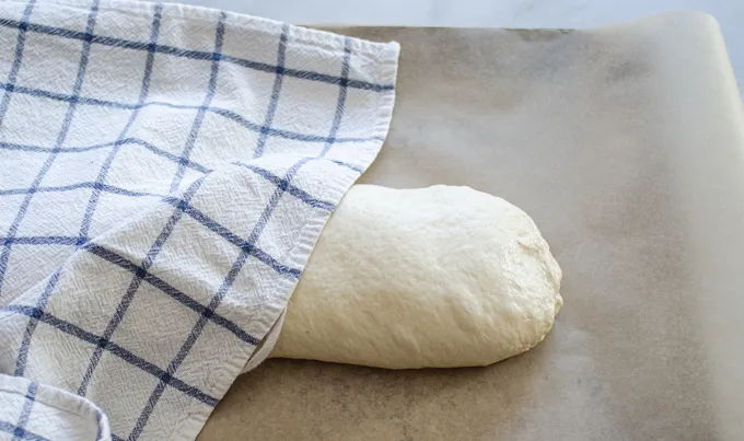 The formed loaf on a parchment lined baking sheet with a tea towel partially covering it.' title='Soft Sourdough French Bread Without Yeast!