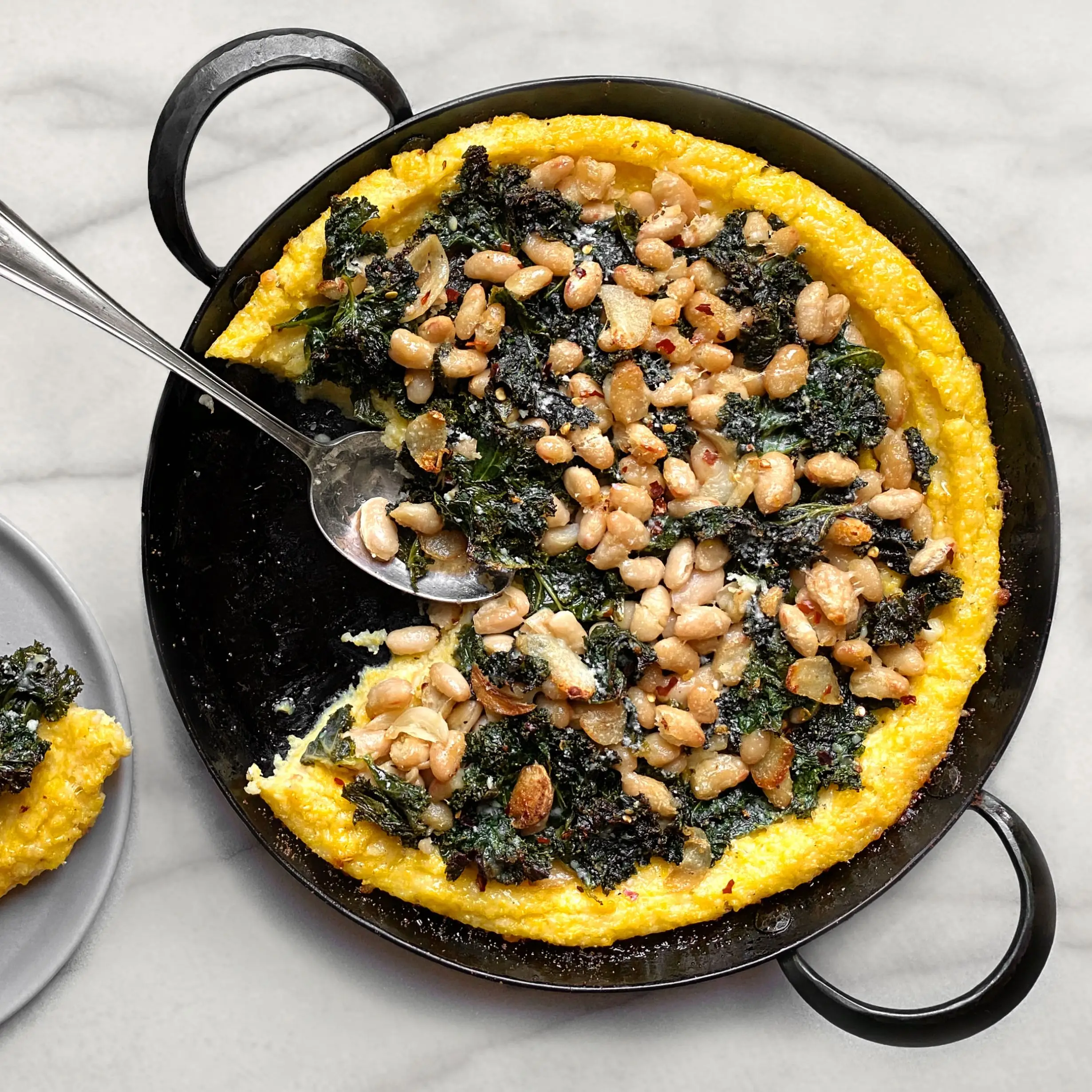 Baked polenta topped with greens and beans in a twohandled pot with a slice of the polenta bake on a plate next to it.