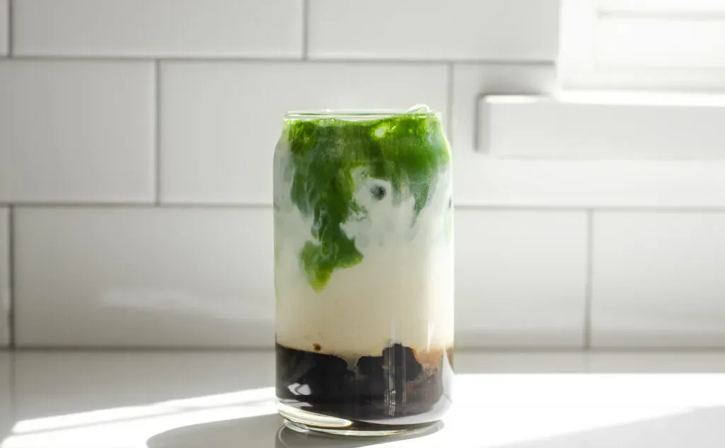 A glass of matcha milk tea on a white countertop in the sunlight.' title='How to Make Matcha Milk Tea with Boba