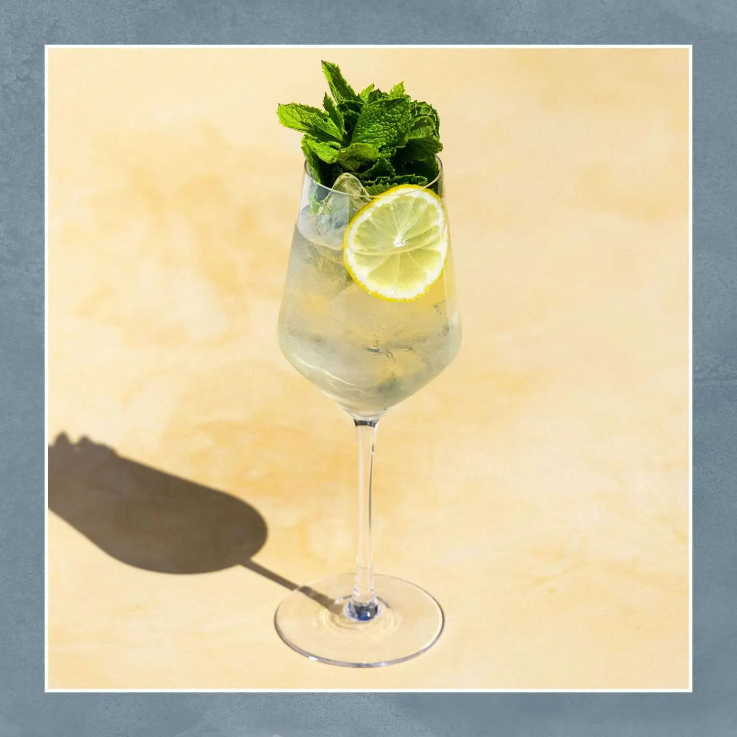 Hugo Spritz cocktail in ice-filled wine glass with lemon wheel and lots of mint, shot on tan background