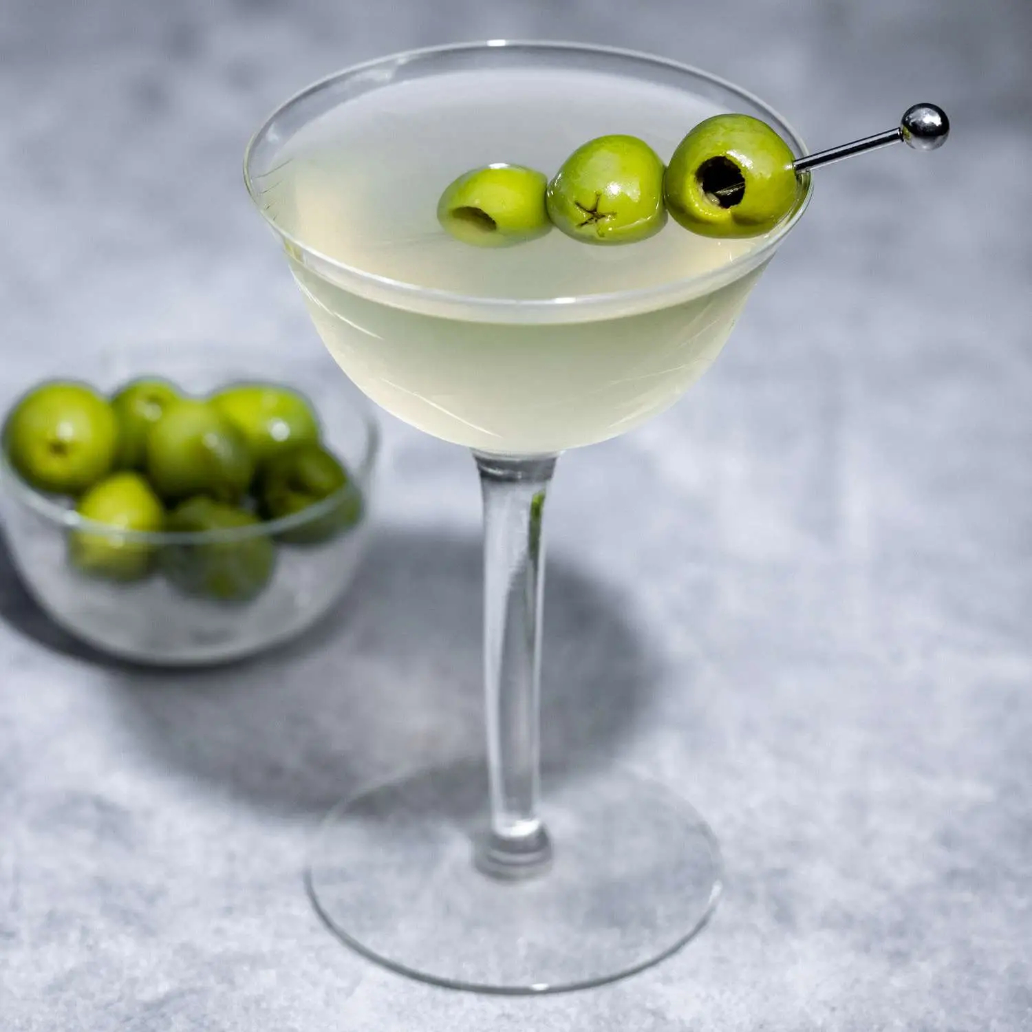 Dirty Martini in a coupe glass with three olives as a garnish, and a side dish of olives in the background
