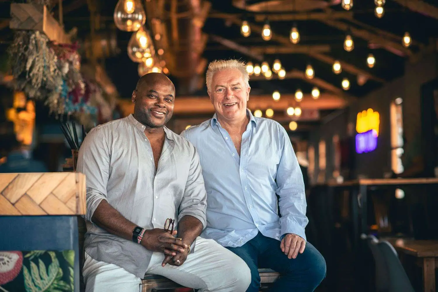 Colin Asare-Appiah and Mark Talbot Holmes seated at bar smiling in button down shirts