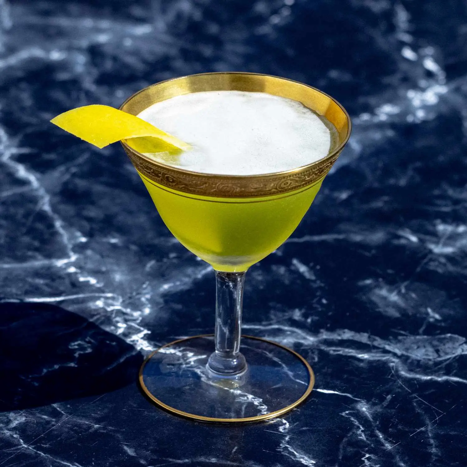 Champs-Elysees cocktail in ornate, gold-rimmed cocktail glass with lemon twist, on dark marble background