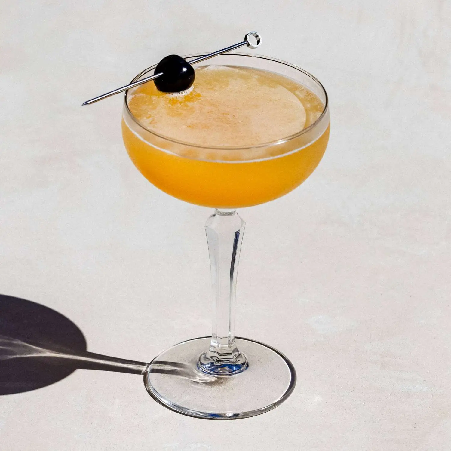 Emerson cocktail, pale orange, in a coupe glass with a maraschino cherry on a metal skewer as garnish