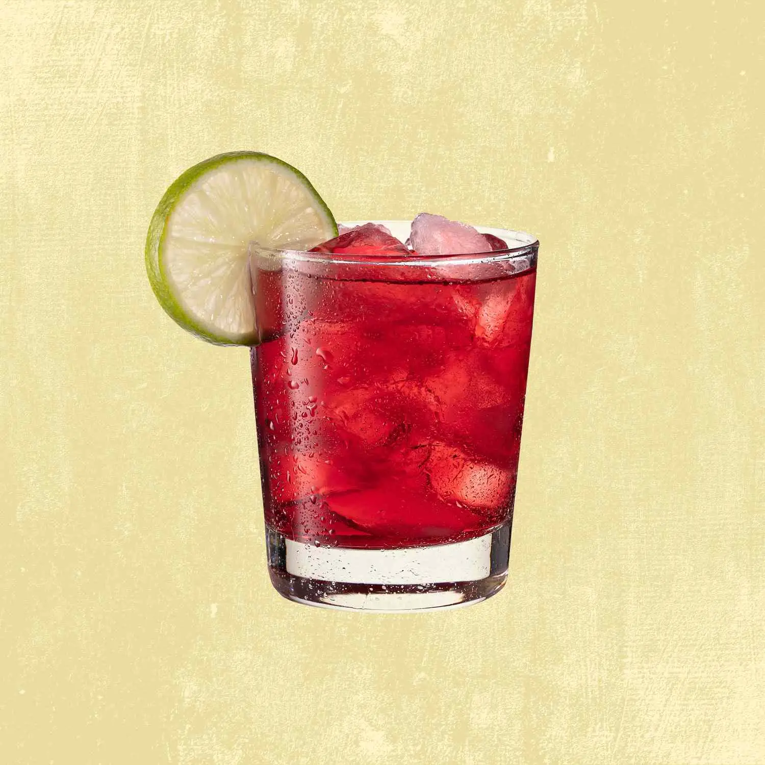 Cranberry and soda in rocks-filled lowball glass with lime wheel against tan background