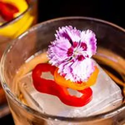 A Little Chili Punch cocktail in a rocks glass with a single large ice cube, and flower and pepper garnish