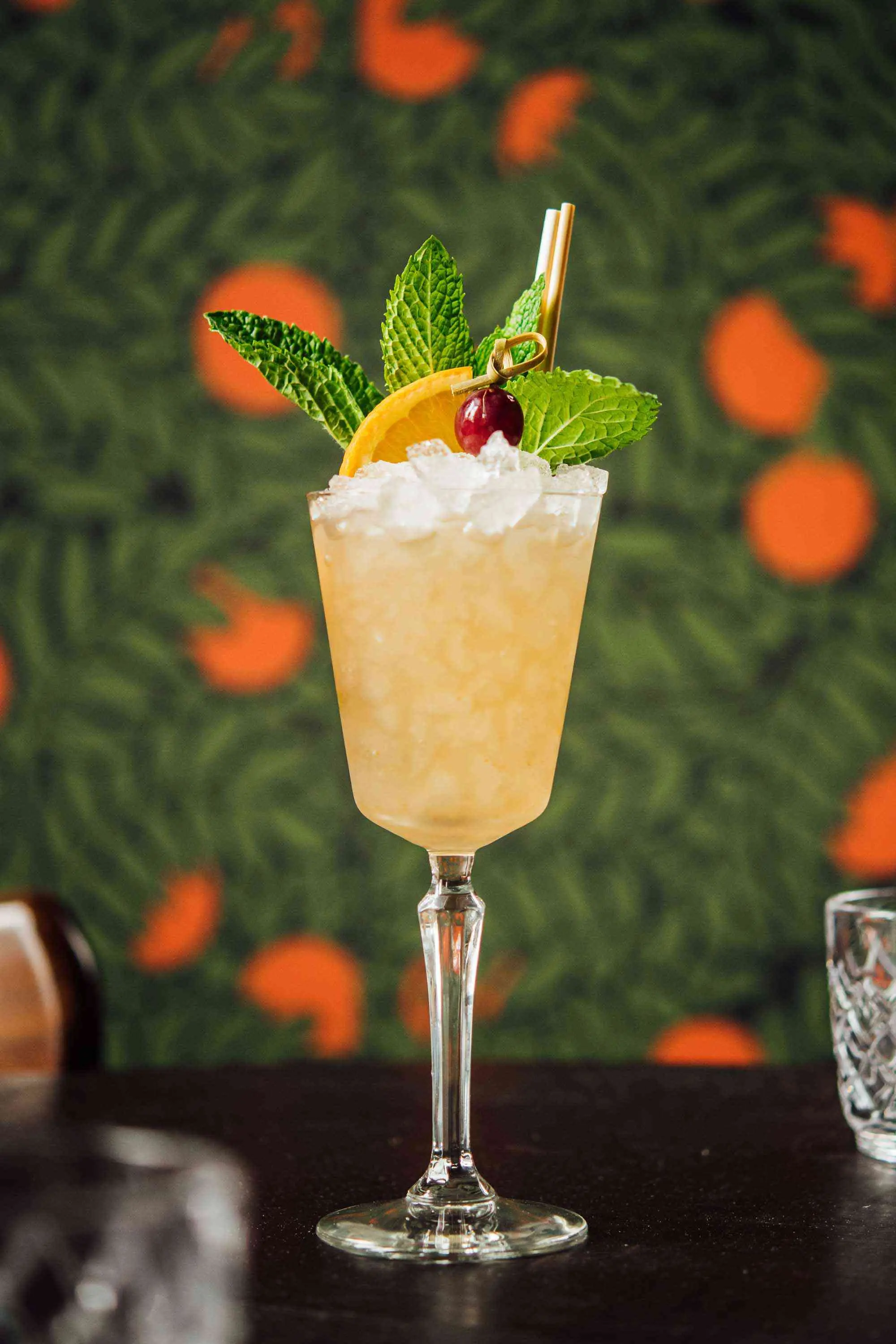 Tropical swizzle style cocktail garnished with mint orange and cherry in front of colorful wallpaper at Jewel of the South