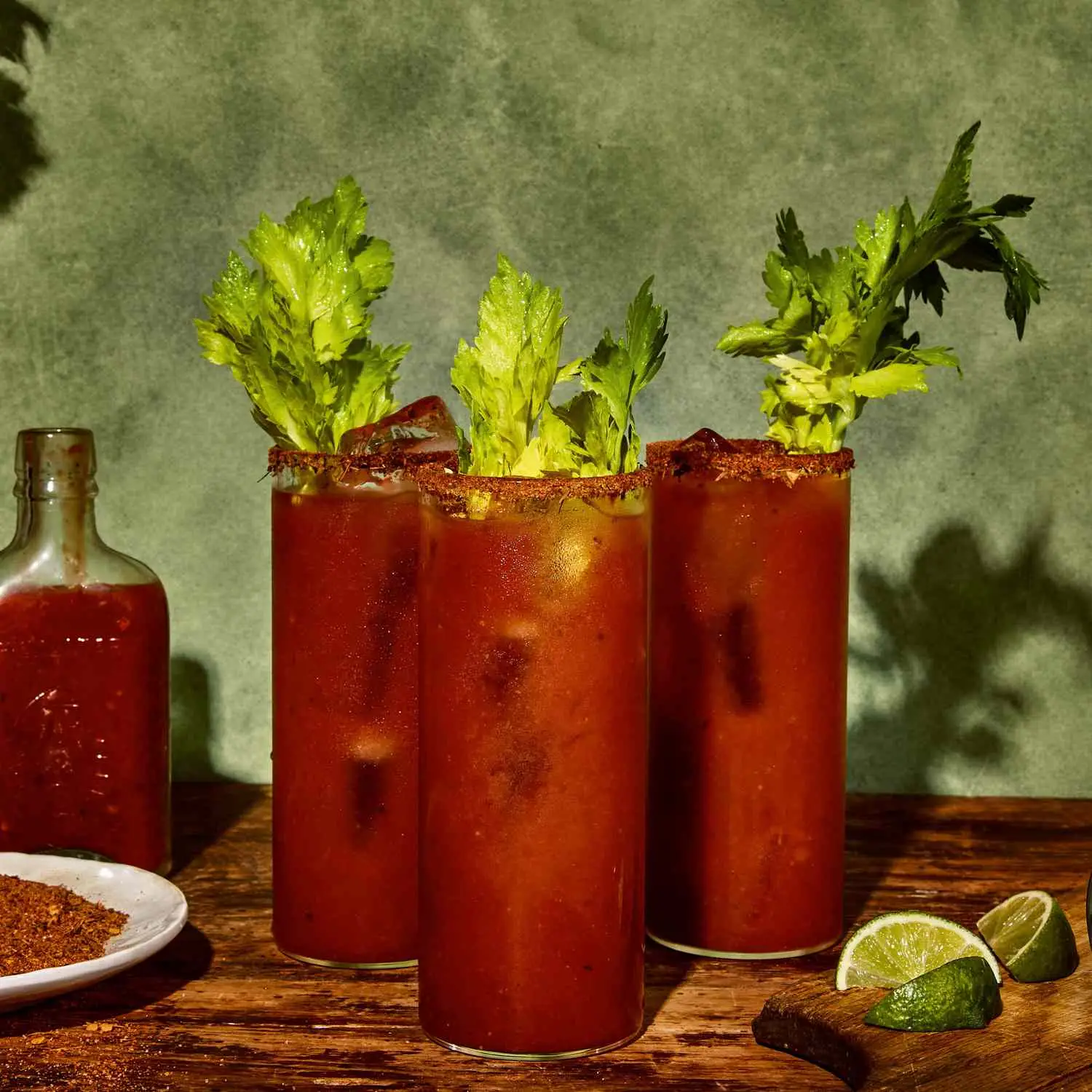 Three Jerk Spiced Bloody Marys in ice-filled highball glasses with jerk spice rim and celery leaf garnish, shot on wooden surface with jerk seasoning, limes, and hot sauce