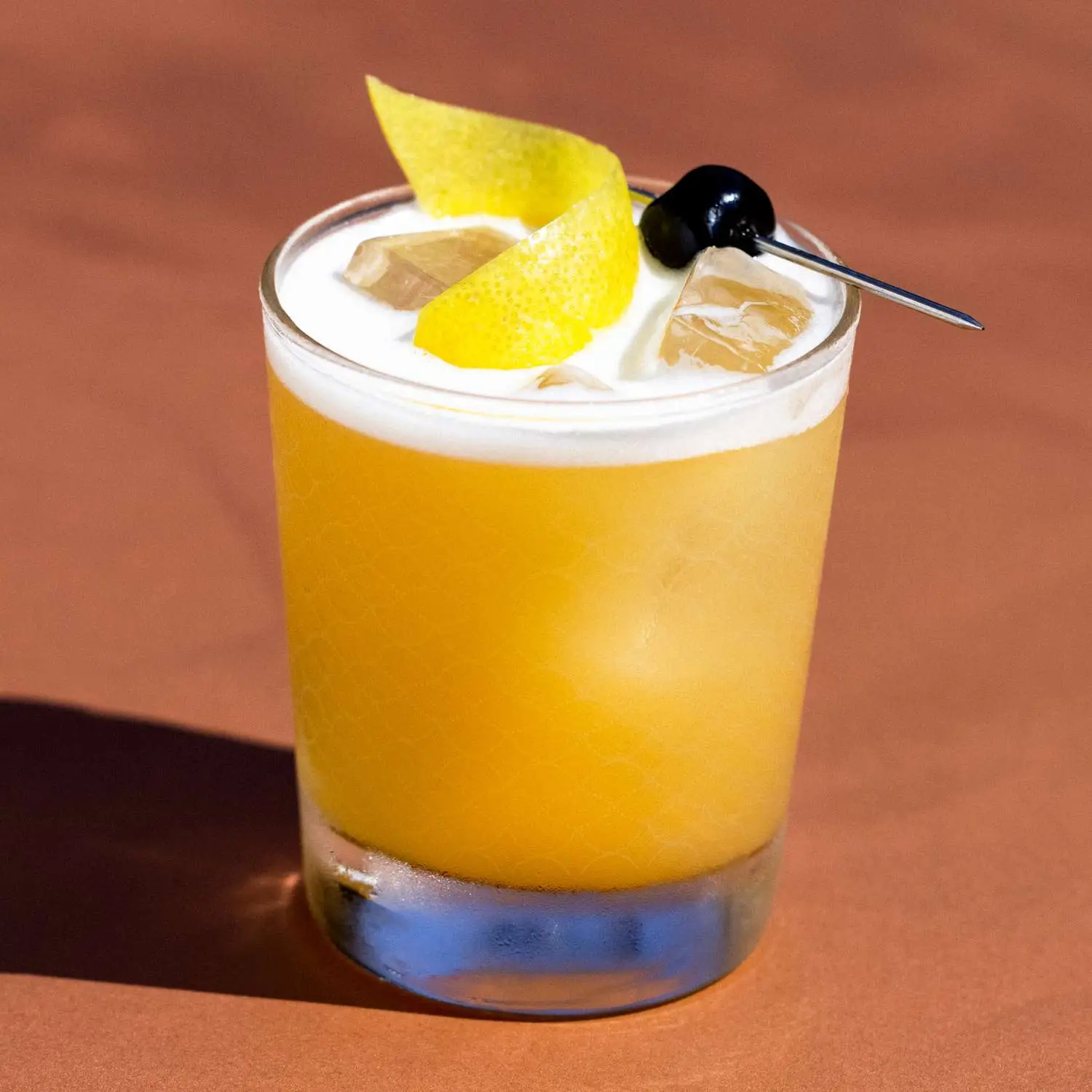 Amaretto Sour in rocks glass with egg white foam on top, garnished with lemon twist and maraschino cherry