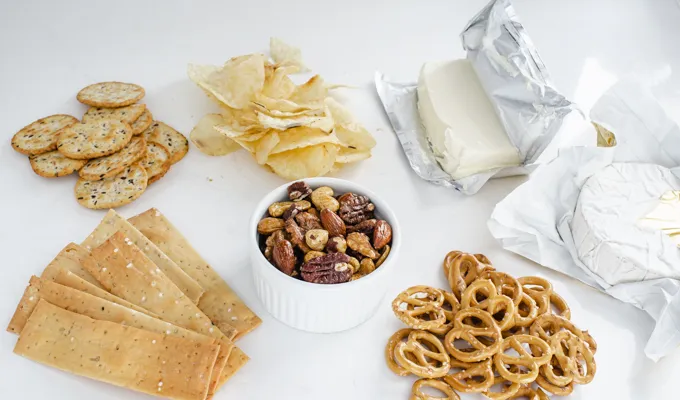 Crackers chips roasted nuts pretzels cream cheese and brie cheese laid out on a counter.' title='Easy Snack Board Ideas