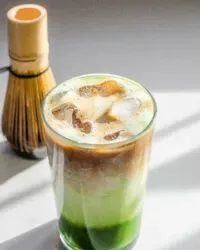 A 12 ounce glass of matcha coffee on a sunny white countertop with a matcha whisk in the background and a white backsplash in the background' title='Matcha Coffee Recipe