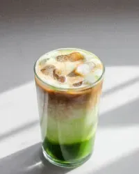 A 12 ounce glass of matcha coffee on a sunny white countertop with a white backsplash in the background' title='Matcha Coffee Recipe