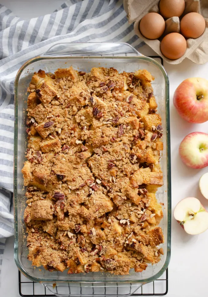 A pan of apple French toast casserole on a wire cooling rack with a striped linen tea towel next to it along with some eggs and apples.' title='Apple French Toast Casserole Recipe