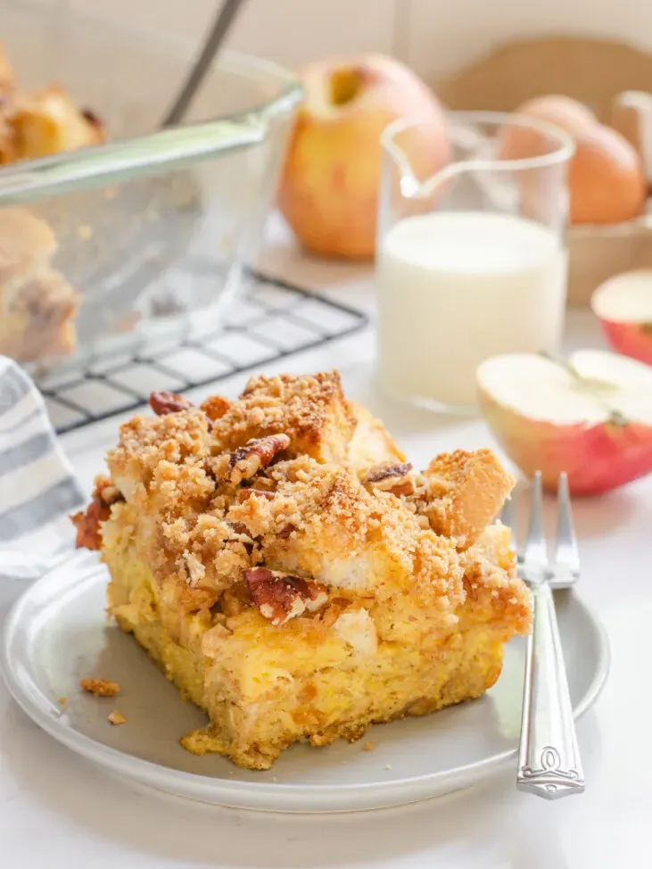 A slice of apple French toast casserole on a grey plate with apples in the background.' title='Apple French Toast Casserole Recipe