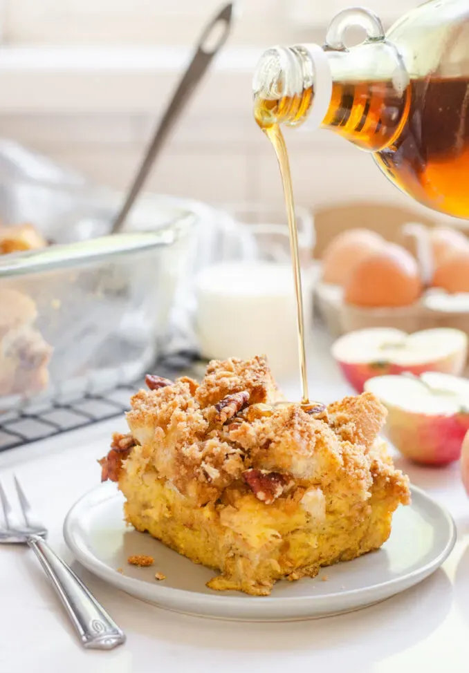 A slice of apple French toast casserole on a grey plate with apples in the background and maple syrup pouring on top.' title='Apple French Toast Casserole Recipe