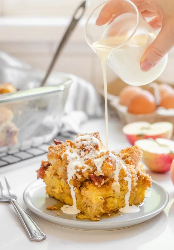 A slice of apple French toast casserole on a grey plate with apples in the background and cream pouring on top.' title='Apple French Toast Casserole Recipe