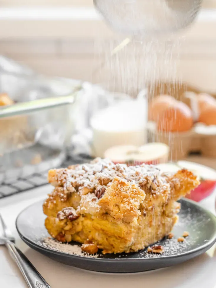 A slice of apple French toast casserole on a grey plate with apples in the background and powdered sugar being sprinkled on top.' title='Apple French Toast Casserole Recipe