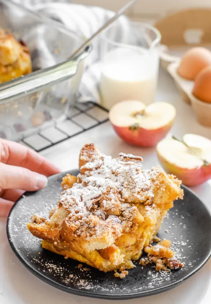 A slice of apple French toast casserole with apples in the background and powdered sugar on top.' title='Apple French Toast Casserole Recipe