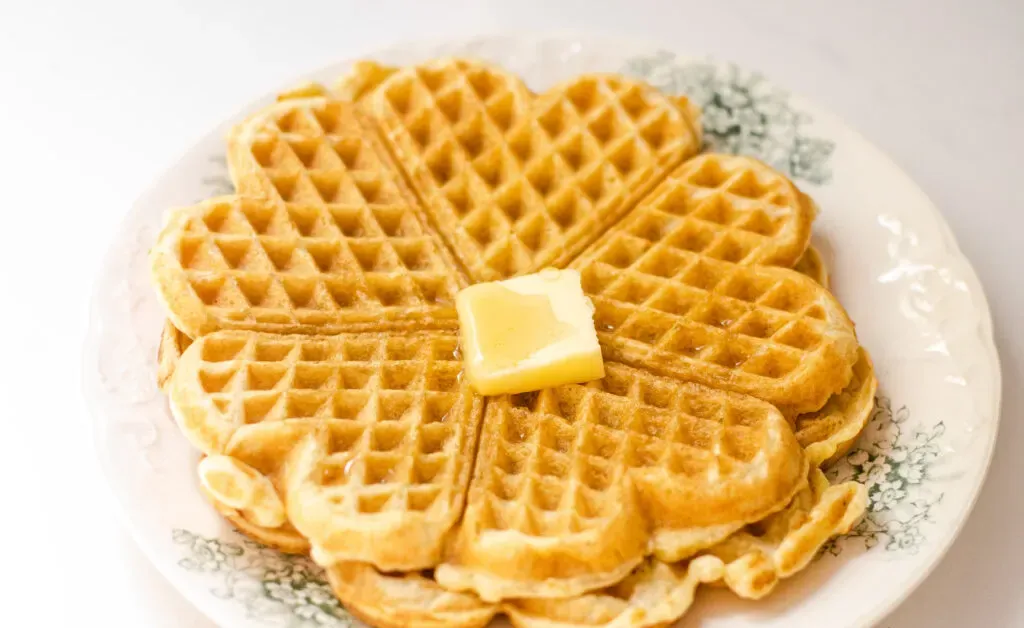A cooked sourdough waffle on a vintage plate with a pat of butter and maple syrup on top.' title='Sourdough Discard Waffles Recipe