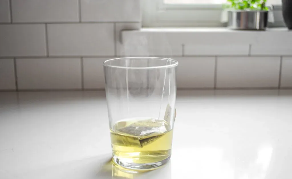 A tea bag and hot water in a drinking glass on a white countertop.' title='Starbucks Peach Green Tea Lemonade Recipe