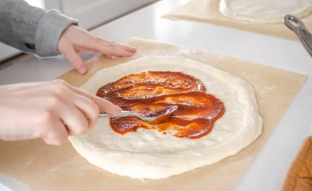 Spreading the pizza sauce on the prepared dough.' title='Sourdough Pizza Crust Recipe