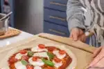 Transferring the topped sourdough pizza crust onto a pizza peel to transfer to the oven.' title='Sourdough Pizza Crust Recipe