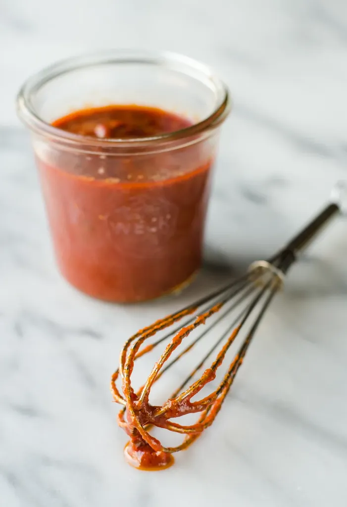 A jar of homemade pizza sauce made with tomato paste on a marble surface with a whisk in front of the jar.' title='Easy Homemade Pizza Sauce with Tomato Paste