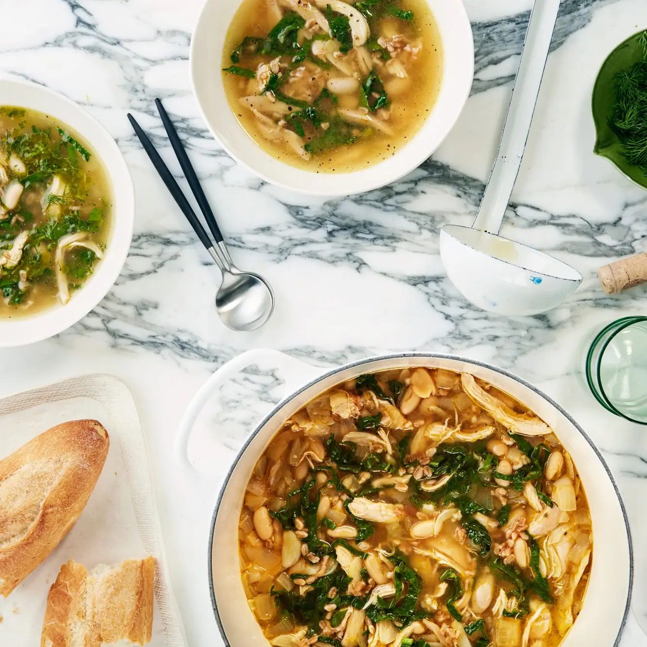 Lemony chicken soup with farro white beans and kale in a white Dutch oven and two bowls.