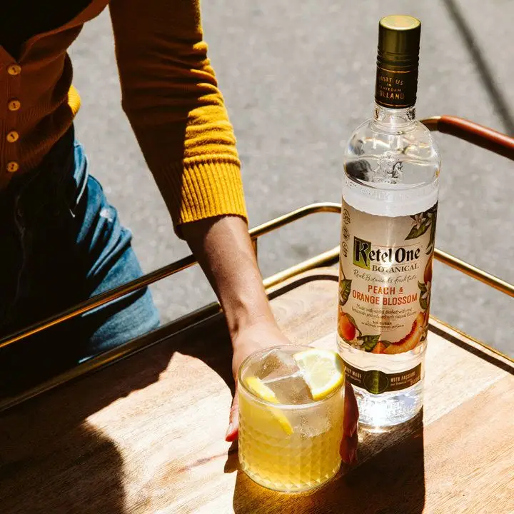 Ketel One Botanical Peach & Orange Blossom vodka bottle on a tray next to a cocktail held by a woman