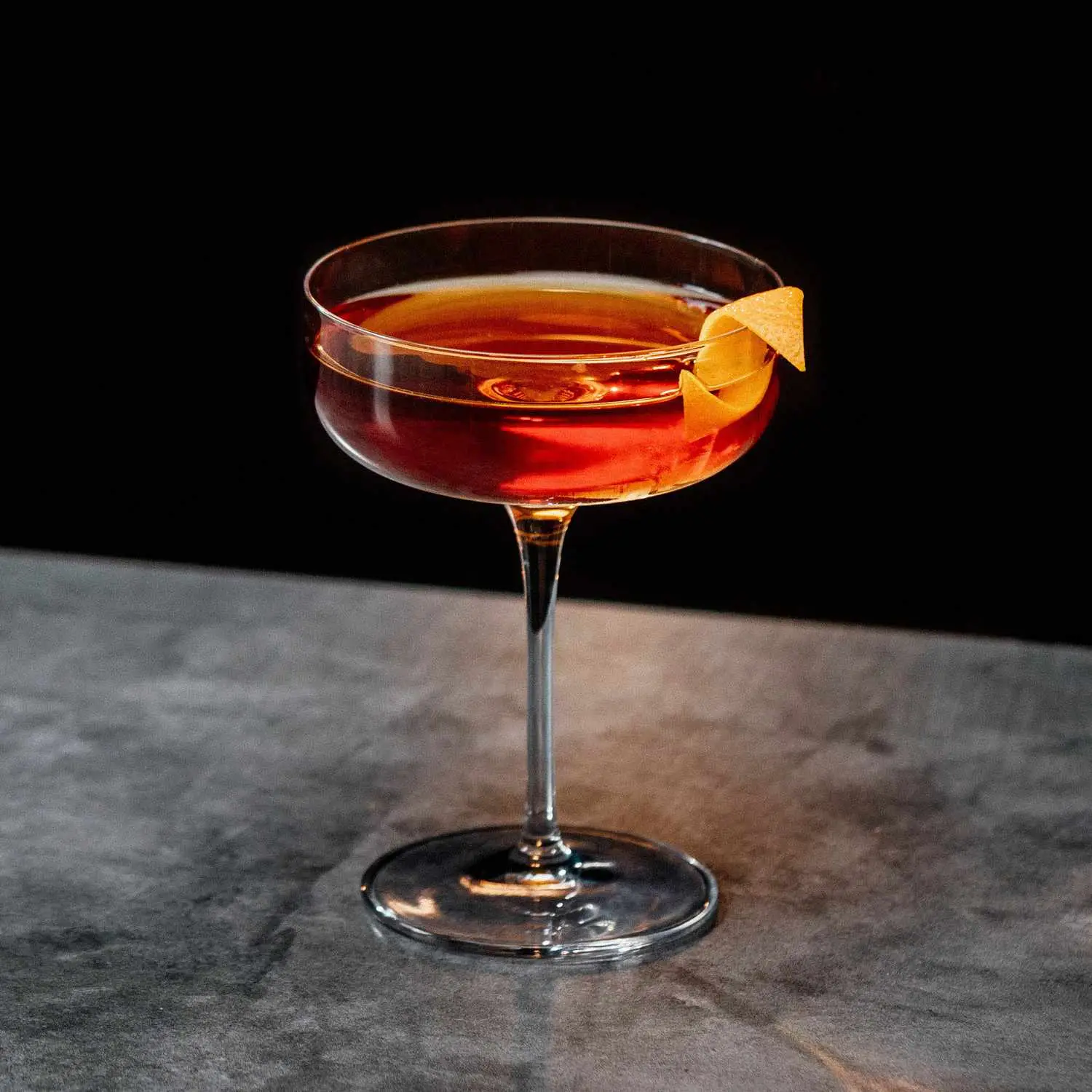 Orange-hued Another Fine Mes cocktail with orange twist in coupe glass, shot on gray surface against black background