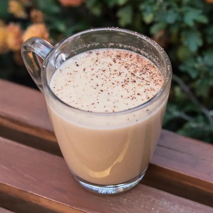 Creamy hot Posset cocktail garnished with nutmeg in glass mug, served outside on slatted wooden table