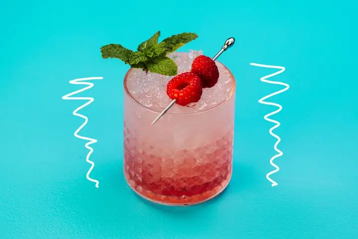 A short, detailed rocks glass holds a bright red drink over crushed ice. The drink is garnished with a sprig of fresh mint and two raspberries pierced on a silver pick. The glass is on a light blue background with a white squiggly line on either side of it. 