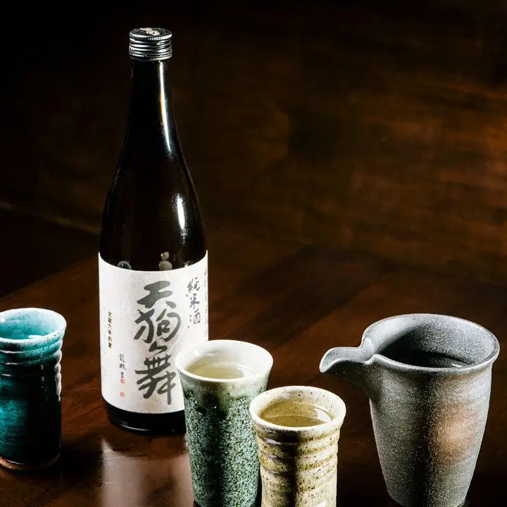 A sake bottle on a dark brown surface surrounded by traditional Japanese ceramic vessels
