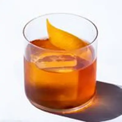 Negroni cocktail with a 3/4 angle view. one large ice cube sits in a thin-glassed tumbler. a large wide angled swath of orange peel sits on the cocktail