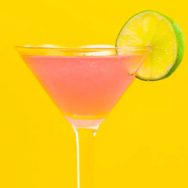 A vivid pink Cosmopolitan in a classic Martini glass is garnished with a whole lime wheel. The background is bright yellow
