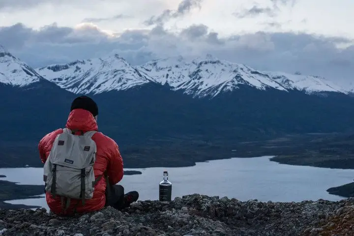 A man in a red jacket sitting next to a bottle of Trakal while looking out over a river and snow-capped mountains