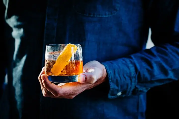 A denim shirt-clad person holding a drink in a rocks glass garnished with an orange twist