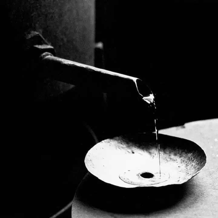 In black and white, crystal clear eau-de-vie pours from a thin spout into a metal dish with a hole in the center. 