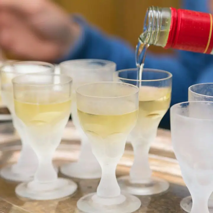 In the foreground are a number of frosty snifters, filled with a yellow-hued absinthe. More absinthe is flowing into one from a red-necked bottle. In the background, out of focus, is an arm in a blue shirt.