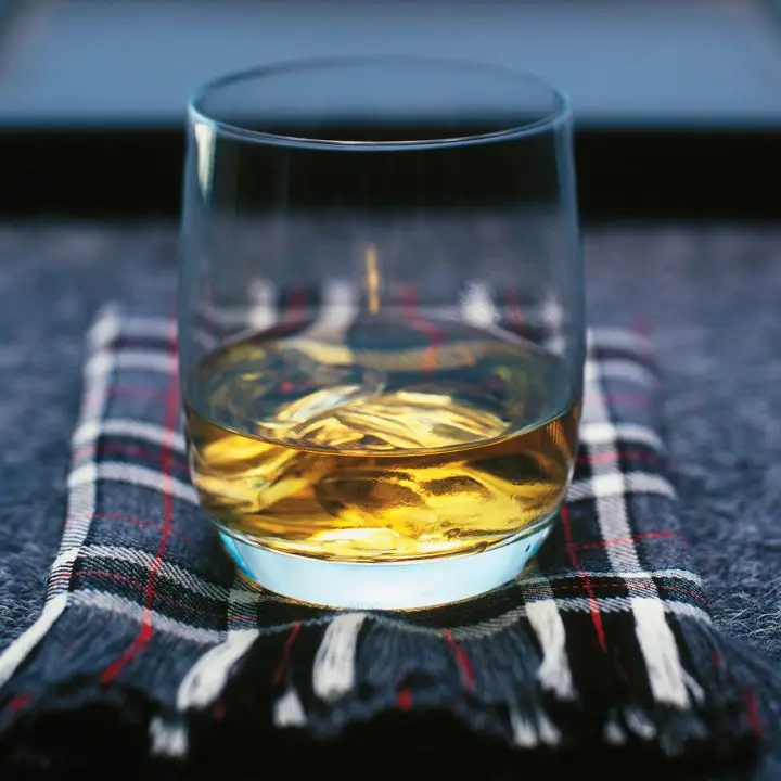 A short, curved glass with a dram of golden Canadian whiskey sits on a tartan scarf. The scarf is patterned with various shades of gray and lines of red. 