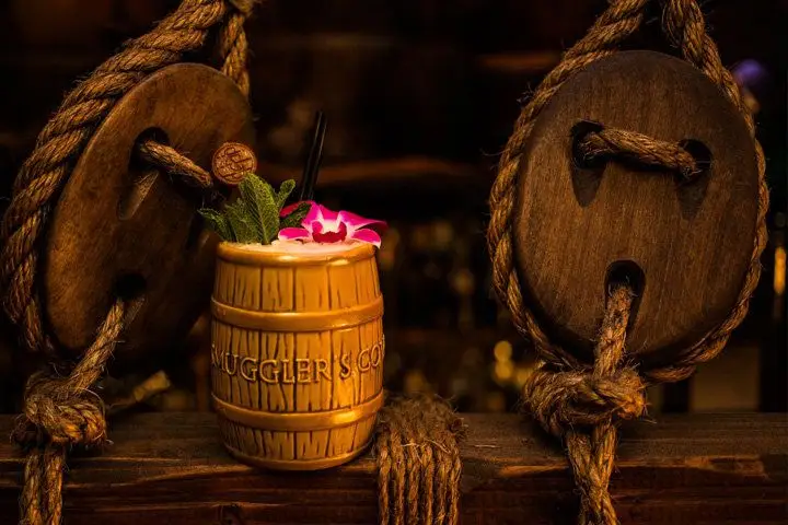 A Smuggler’s Cove-branded tiki mug garnished with a flower and mint sprig set atop a wooden beam accented with rope