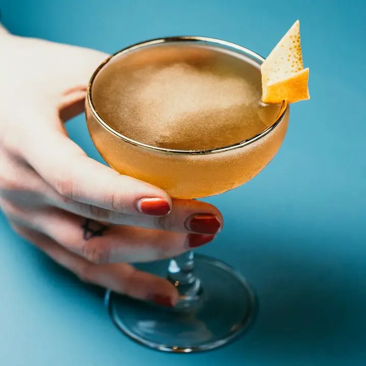 A hand with red fingernails holding a shaken drink in a coupe glass garnished with an orange twist