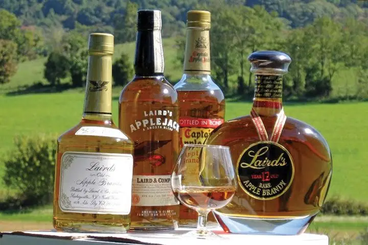 A variety of different styles and shapes of Laird’s Apple Brandy bottles and a snifter full of said brandy sit on a table outside on a sunny day. In the background there’s a swooping hill topped with a copse of trees.