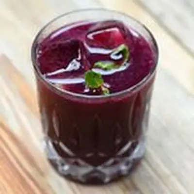 A faceted rocks glass holds a bright purple drink garnished with a few small mint leaves. The glass is resting on a wooden surface.
