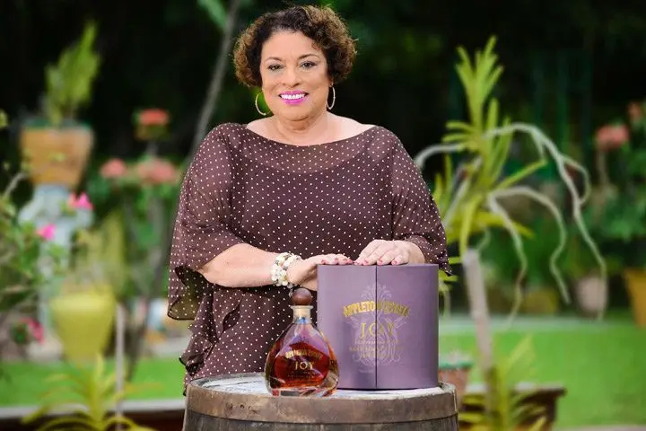 A woman in a dark purple polka-dotted top stands smiling behind a bottle of Appleton Estate and its violet-hued box