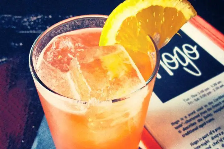 A bright orange cocktail on ice garnished with an orange slice next to an orange, black, and white menu