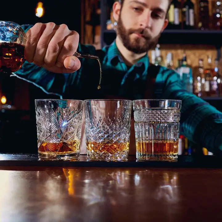 A bearded bartender in a blue plaid shirt pours a stream of Irish whiskey into a patterned rocks glass. Two other glasses flank the center one that he is pouring into, each with a large dram of whiskey.