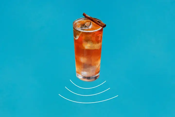 On a blue background, a tall collins glass is filled with eye and a spritzy red highball. A cinnamon stick rests on the lip of the glass