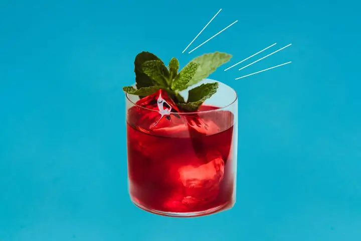 On a sky blue background, a short, clean cut rocks glass is filled with ice and a berry-red drink, garnished with a sprig of fresh green mint